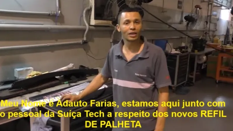 Um homem de camisa cinza fala para a câmera, ao lado de uma bancada com palhetas, numa área de serviço com ferramentas ao fundo.